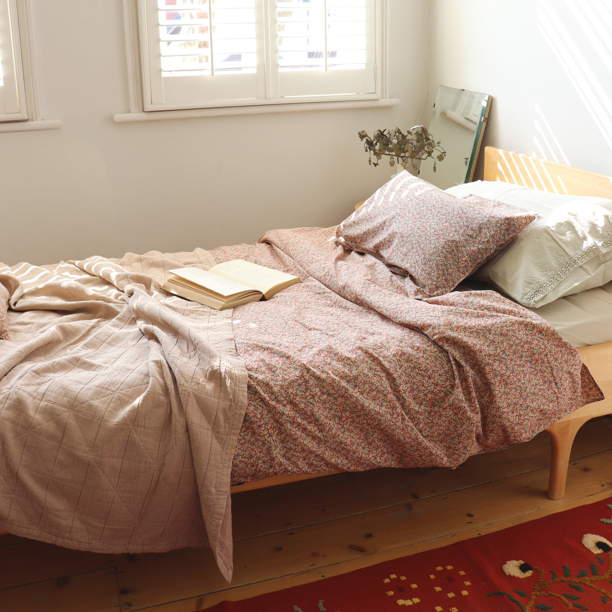 A cosy bed with camomile london floral printed bedding in soft pinks and red colours with rose pink cotton textured blanket at bottom of bed in a cream room with soft sunlight.