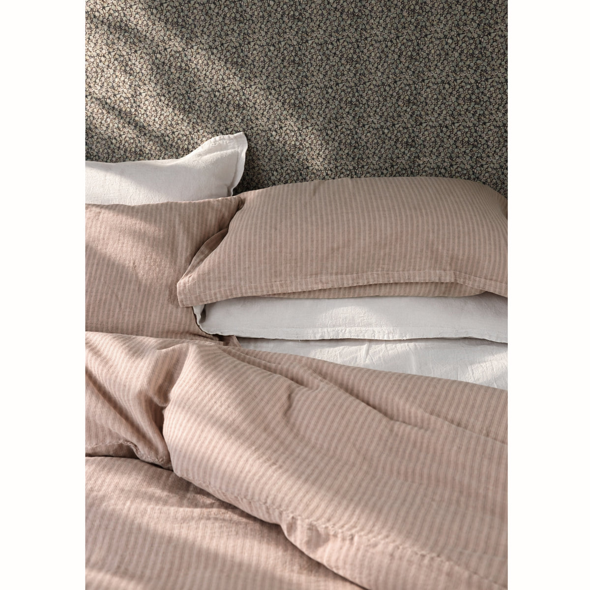 A close-up of a bed made with a soft rose pink and taupe vertically striped duvet cover and matching pillowcases, layered over off white sheets, against a dark floral headboard.